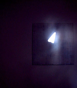 Ein sehr dunkles Zimmer. Man sieht eine Wand, an der ein Bild zu erkennen ist, auf der ein helles Dreieck zu erkennen ist.